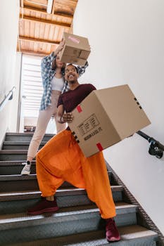 A playful interracial couple carrying cardboard boxes down the stairs during a move-in.