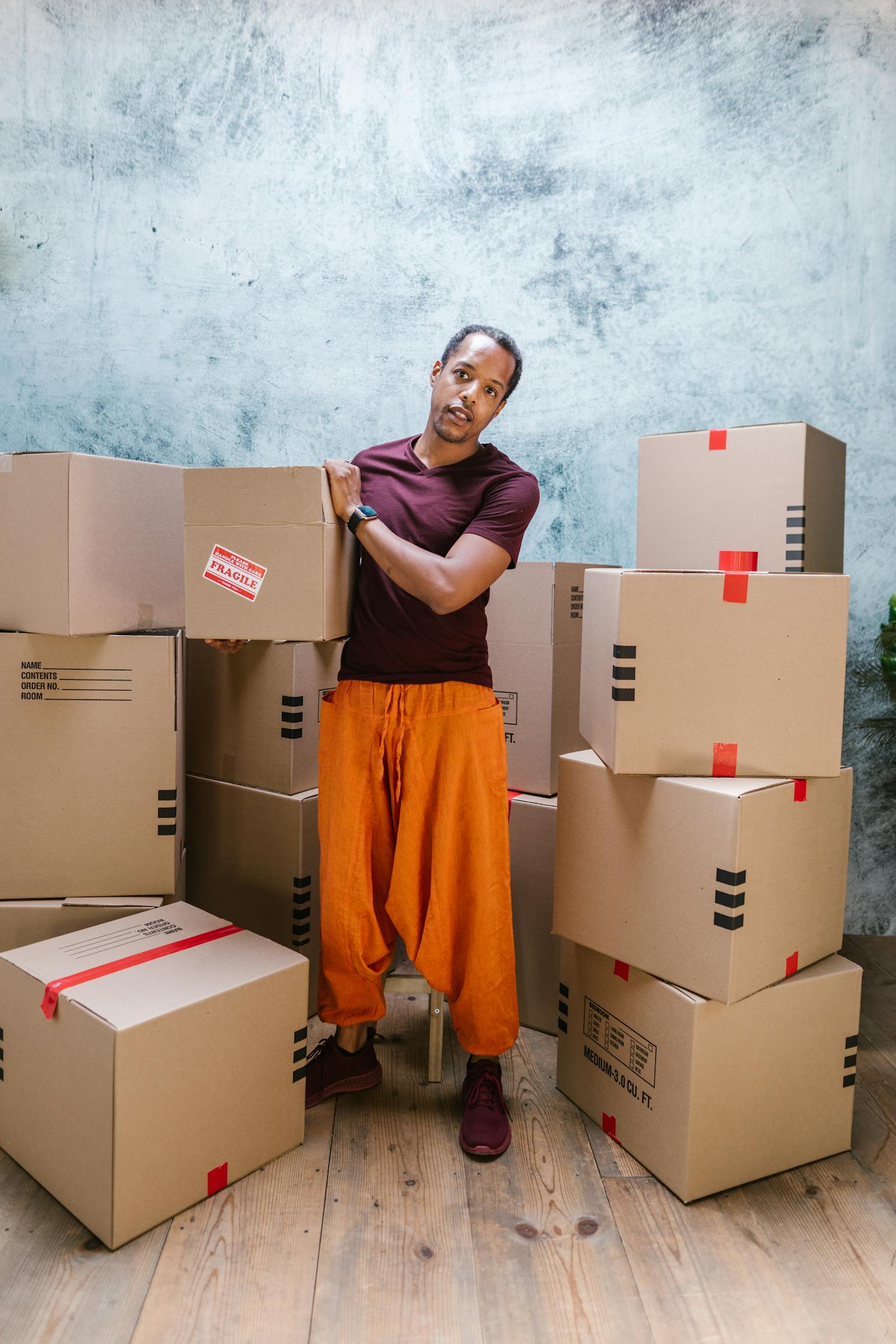 Man in Orange Pants Holding a Package Standing Beside Cardboard Boxes