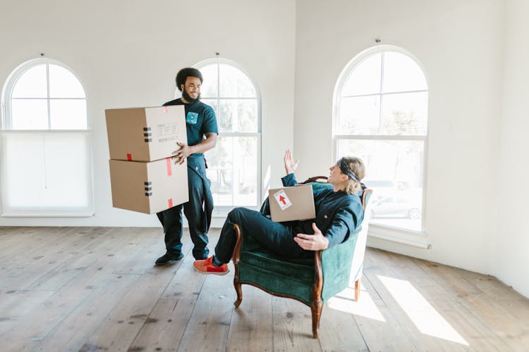 A Man Standing Carrying Boxes Talking To A Man Sitting On A Chair