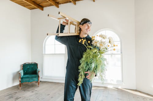 Man in a Jumpsuit Wearing a Bandana Carrying a Wooden Stool and Flowers in a Vase