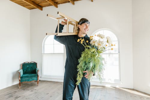 Free Man in a Jumpsuit Wearing a Bandana Carrying a Wooden Stool and Flowers in a Vase Stock Photo