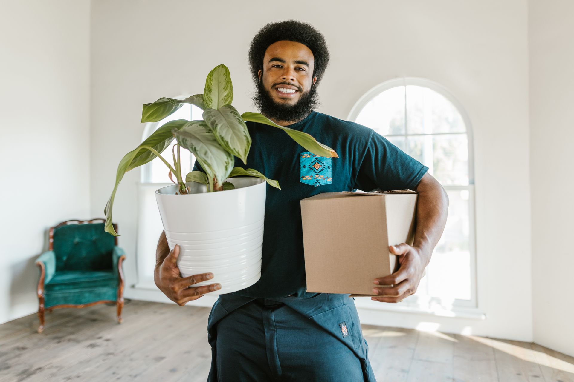 African American man holding a potted plant and box while moving into a new house.