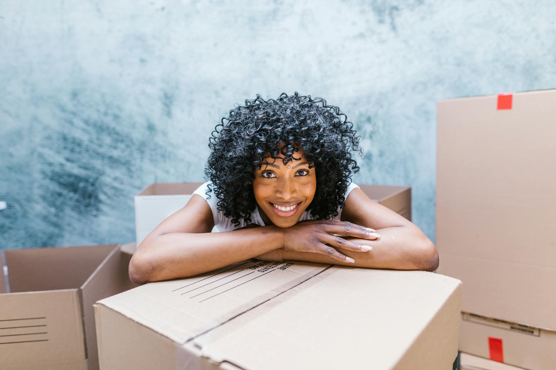 Happy woman with curly hair leaning on moving boxes indoors, ready for relocation.