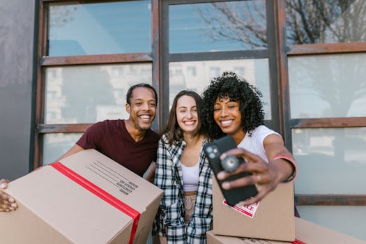 Three friends taking a selfie while moving into a new home with boxes around them.