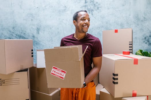 Happy man in maroon shirt holding a fragile box among packages indoors.