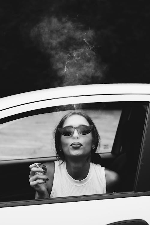 Grayscale Photo of a Woman in a Car Smoking a Cigarette