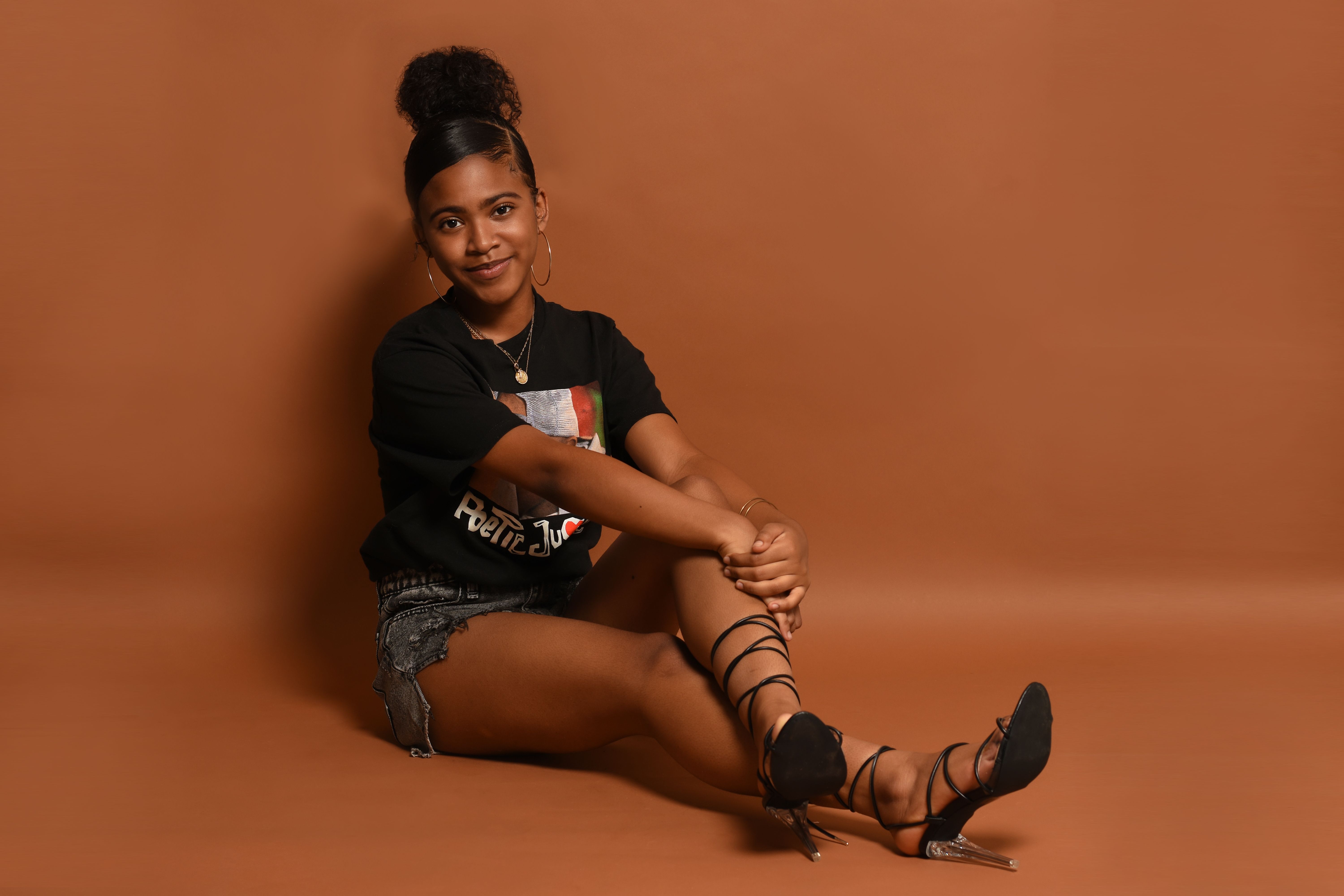 Stylish African American woman in black tee and shorts, sitting on brown background.
