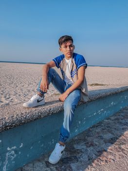 Casual young man in jeans and sneakers sitting on a beach wall with clear blue sky.