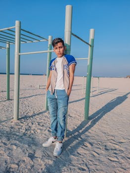 Man in casual wear poses by monkey bars at a sandy beach playground under clear skies.