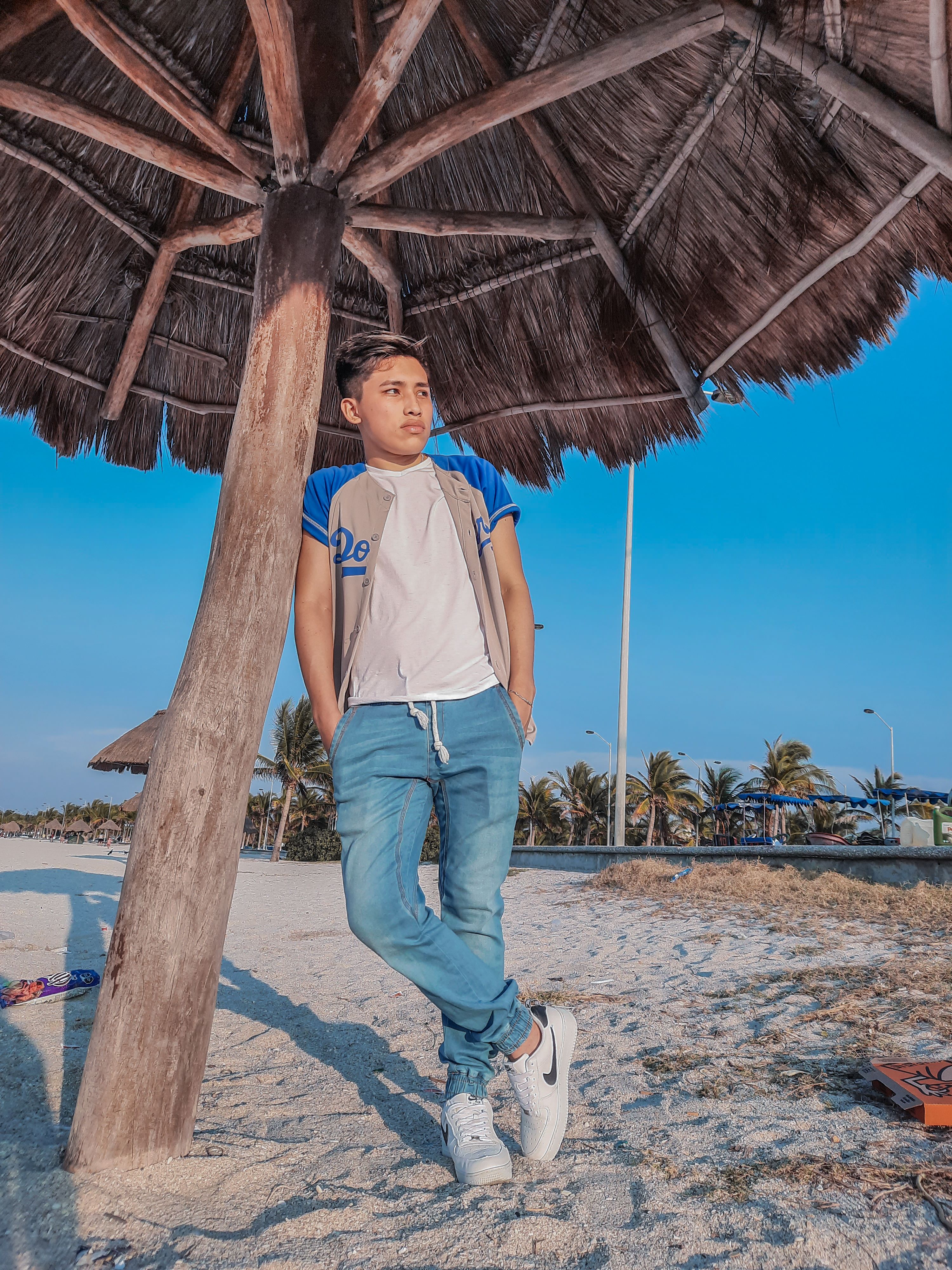 A young man stands casually under a wooden beach umbrella on a sunny day.