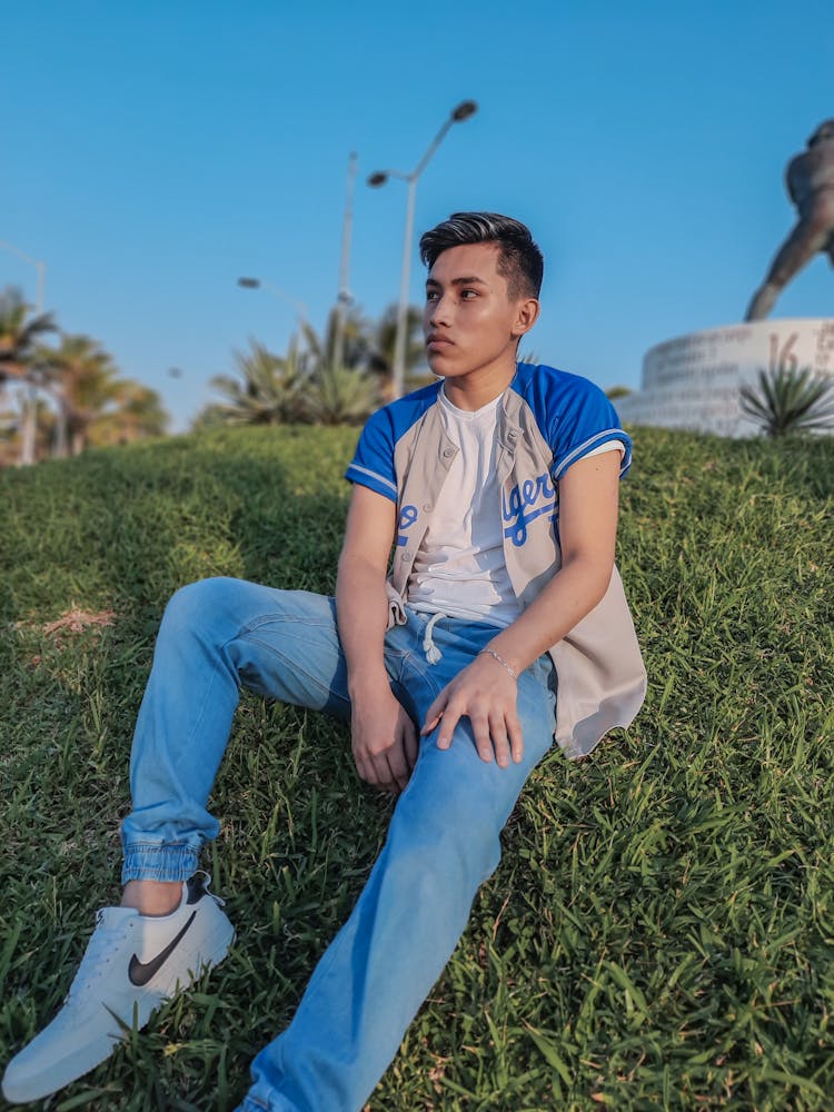 A Boy In A Gray And Blue Jersey Looking Away While Sitting On Green Grass