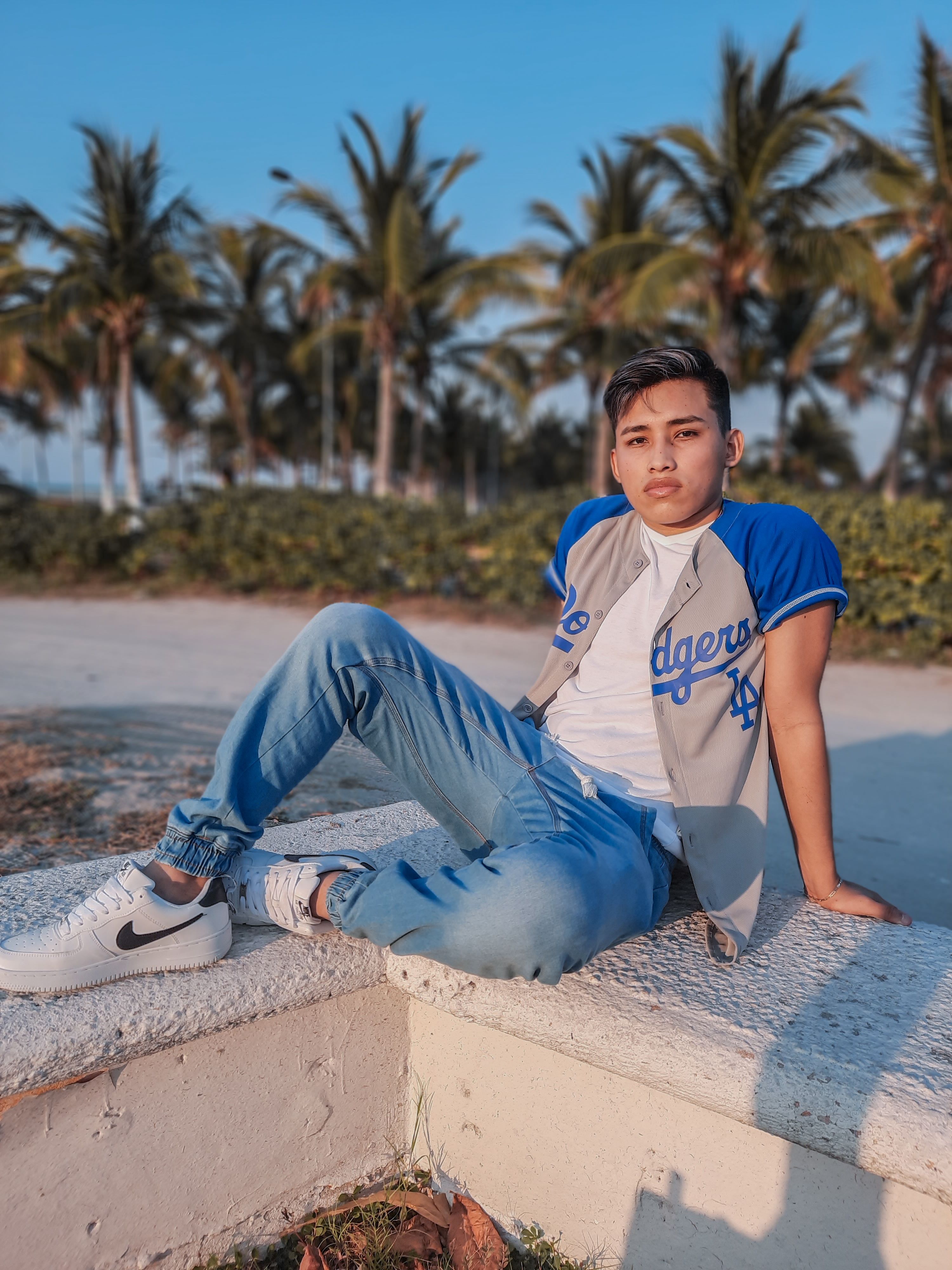A young man poses stylishly on a beach, relaxing under palm trees in casual attire.