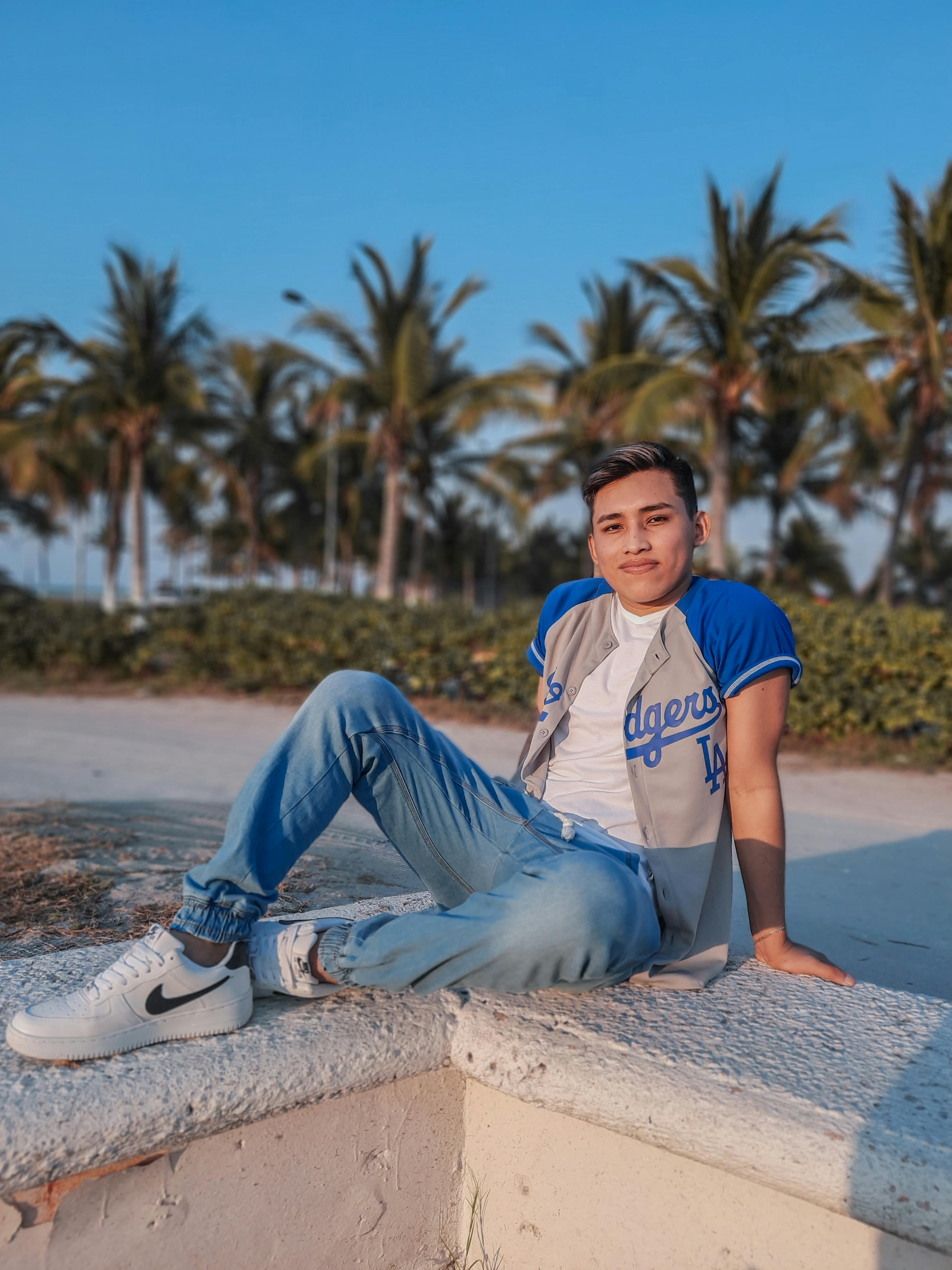 Casual young man sitting on a wall by a tropical beach, smiling in the sun.