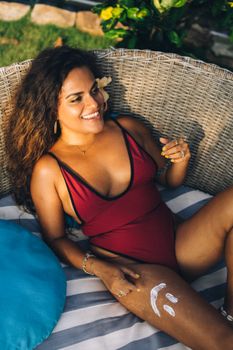Smiling woman in swimsuit relaxing on a wicker chair with sunshine and greenery.