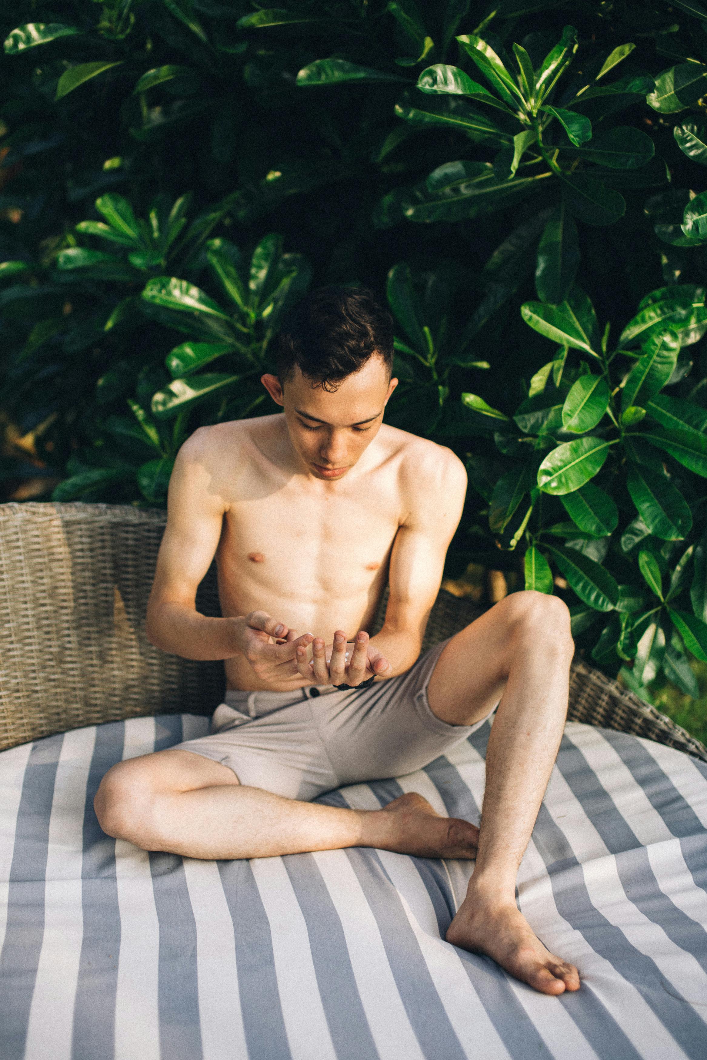 Shirtless man sitting in the sun, applying sunscreen for skin protection outdoors.
