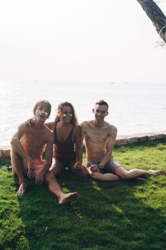 Three friends in swimwear sitting on grass by the ocean, smiling and enjoying the sun.