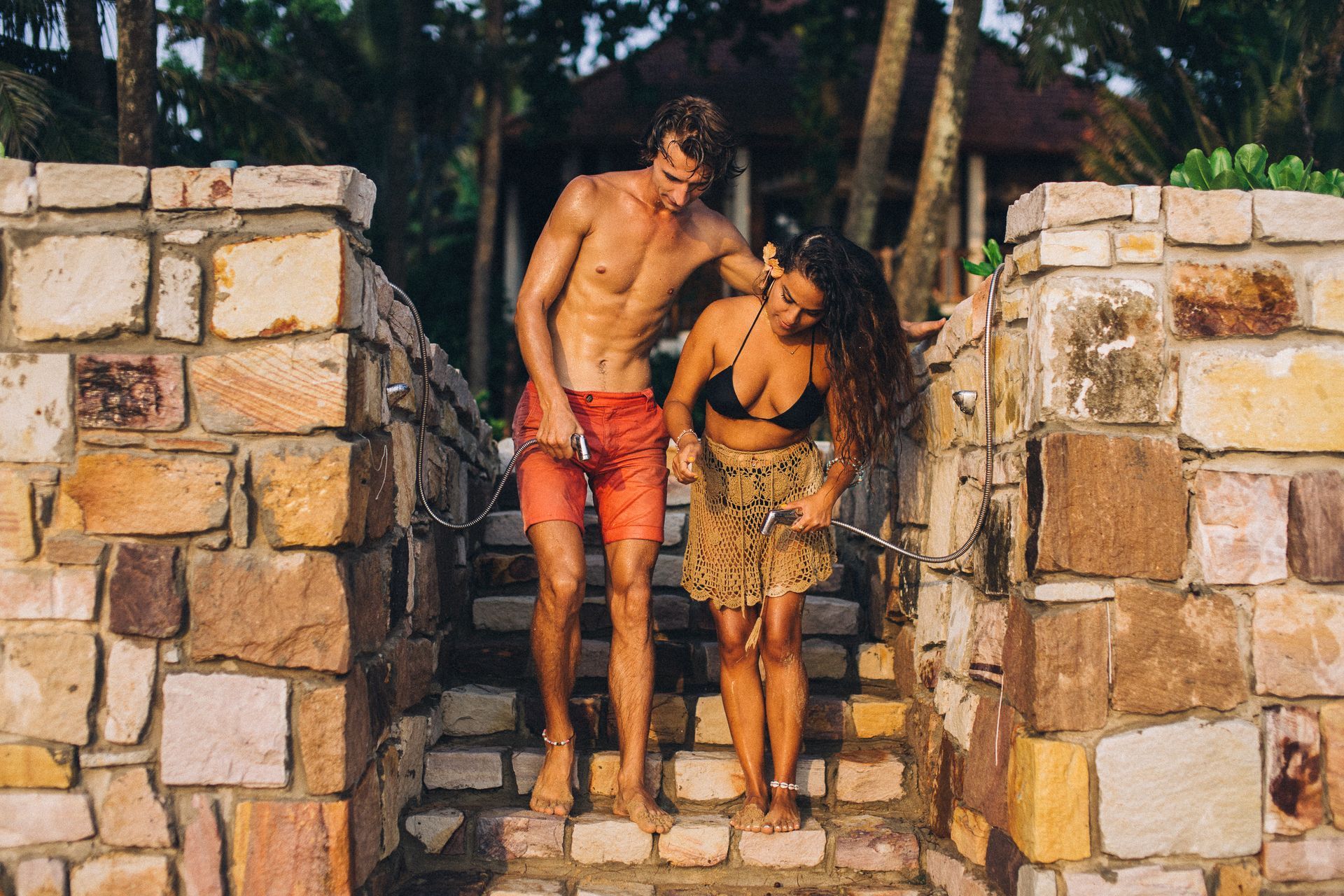 A couple in swimwear rinsing off at an outdoor shower surrounded by stone walls.