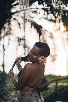 Smiling woman embraces refreshing outdoor shower in a lush, tropical environment.