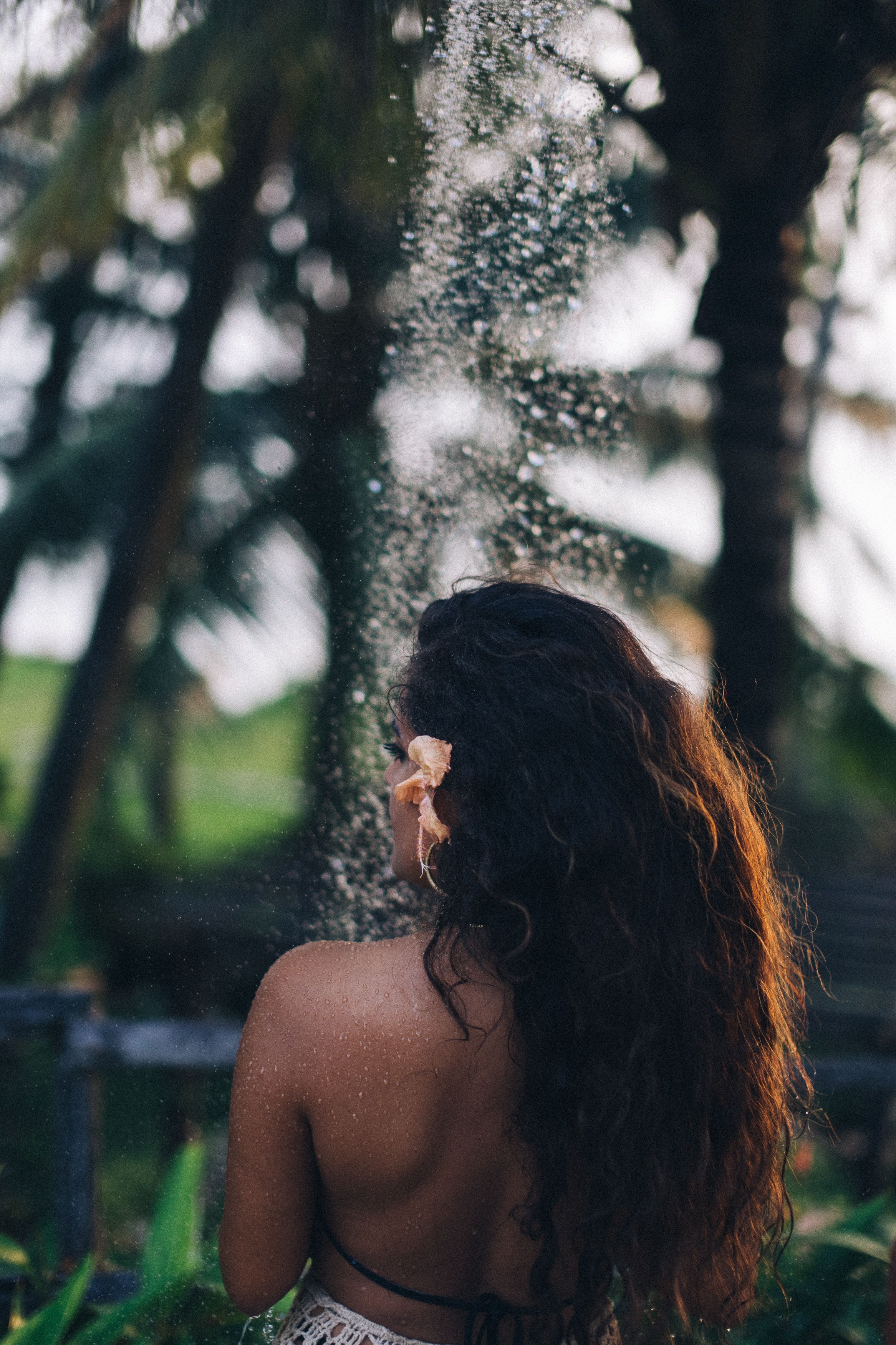 A woman with long hair enjoys an outdoor shower in a tropical setting with palm trees.