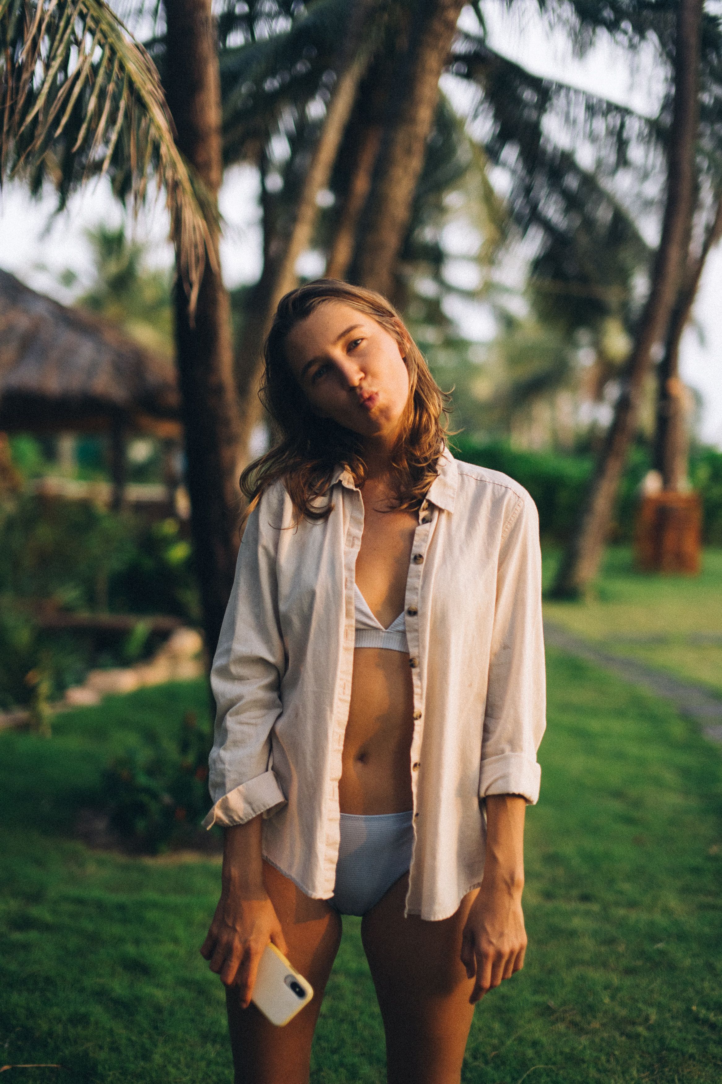 Young woman in a bikini holding a phone, posing playfully in a tropical outdoor setting.