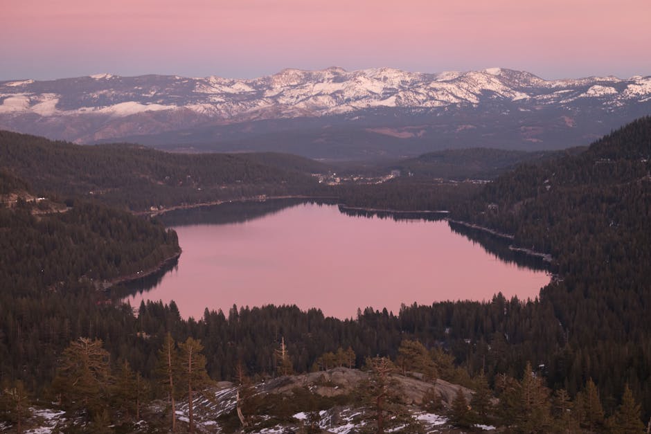 Breathtaking view of a mountain lake reflecting a pink sky at sunset, surrounded by snow-capped peaks and dense forests.