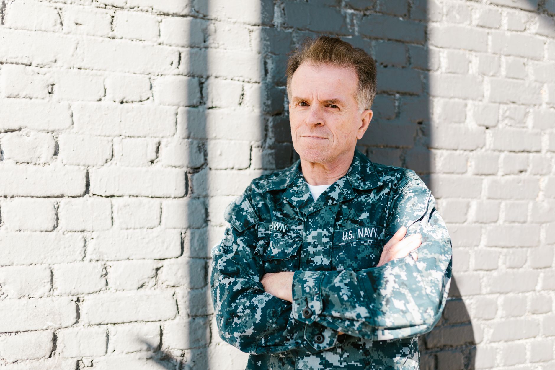 Senior Navy veteran standing proudly in naval uniform against a brick wall, outdoors in daylight.