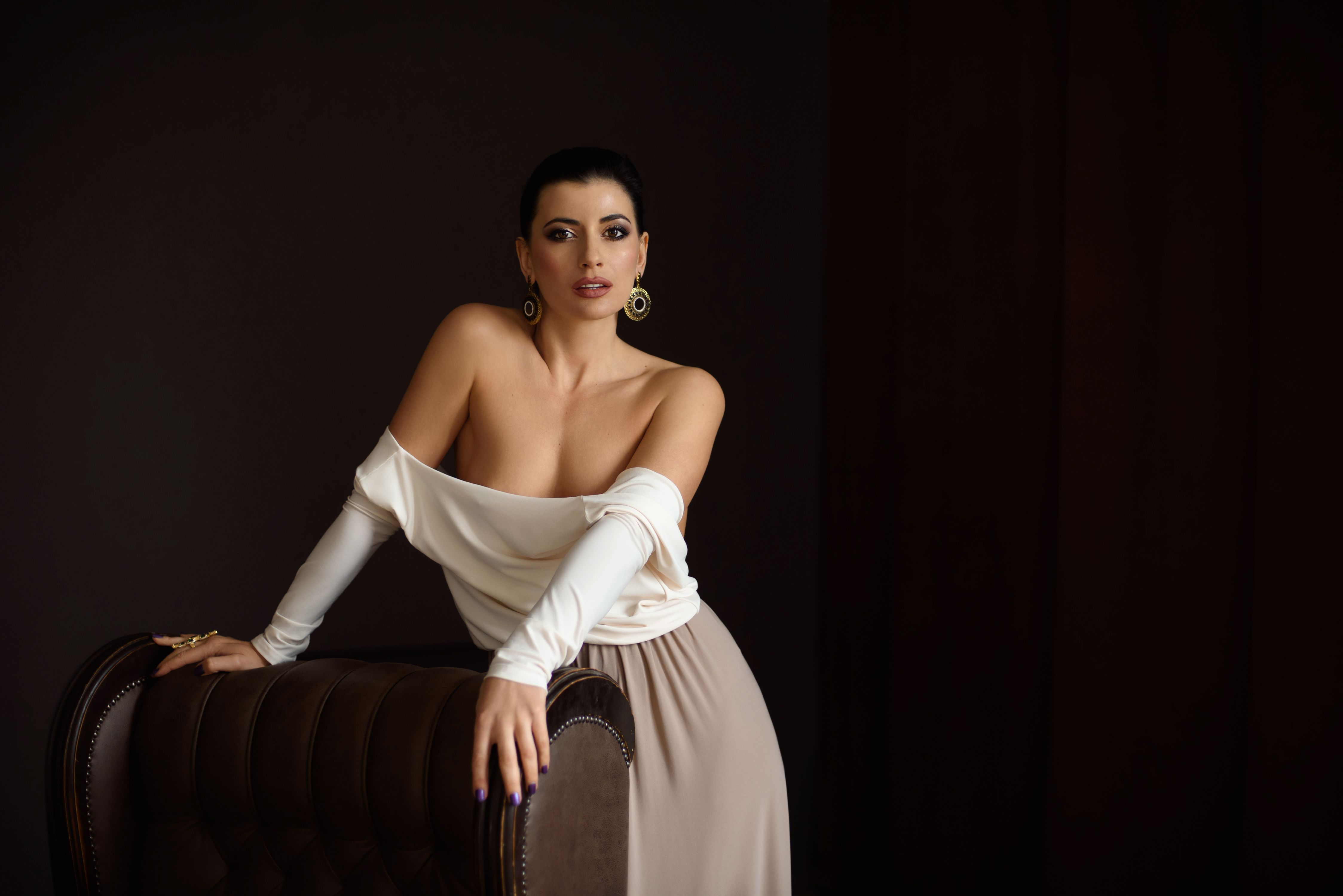 Gorgeous female in stylish outfit with bare shoulders and with earrings standing near armchair and looking at camera on dark background