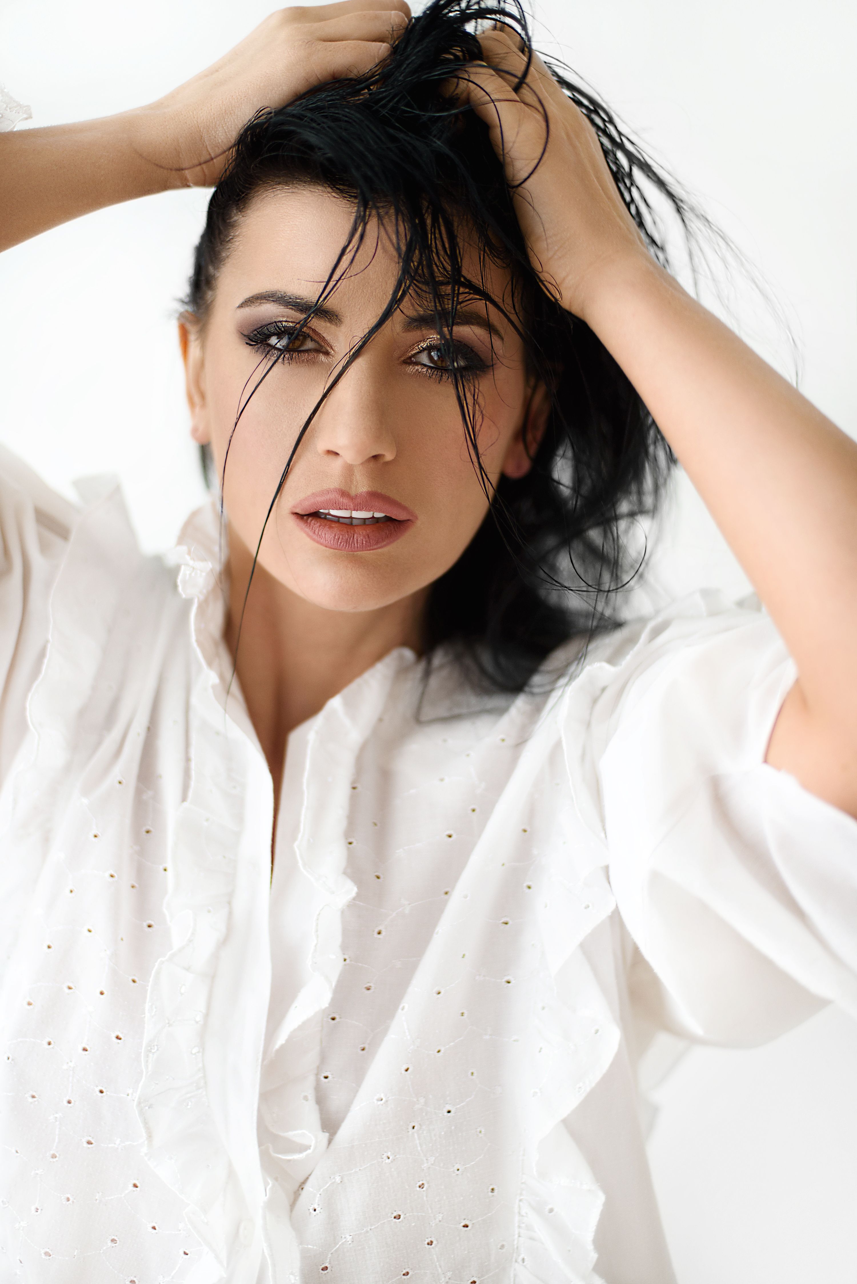 Charming female in white shirt tousling dark hair and looking at camera while standing on white background in light studio