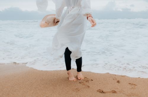 Free Unrecognizable person in a white dress walking along a sandy shore, holding a hat. Stock Photo