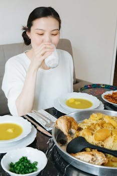 Asian woman drinking at a table set with soup, chicken, and rice dishes in an indoor setting.