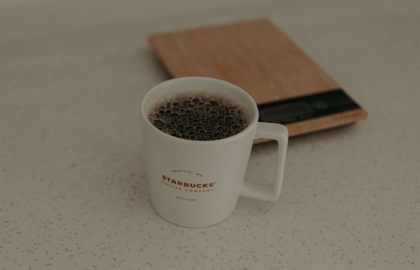 A close-up of a Starbucks coffee mug filled with coffee next to a weighing scale on a speckled surface.