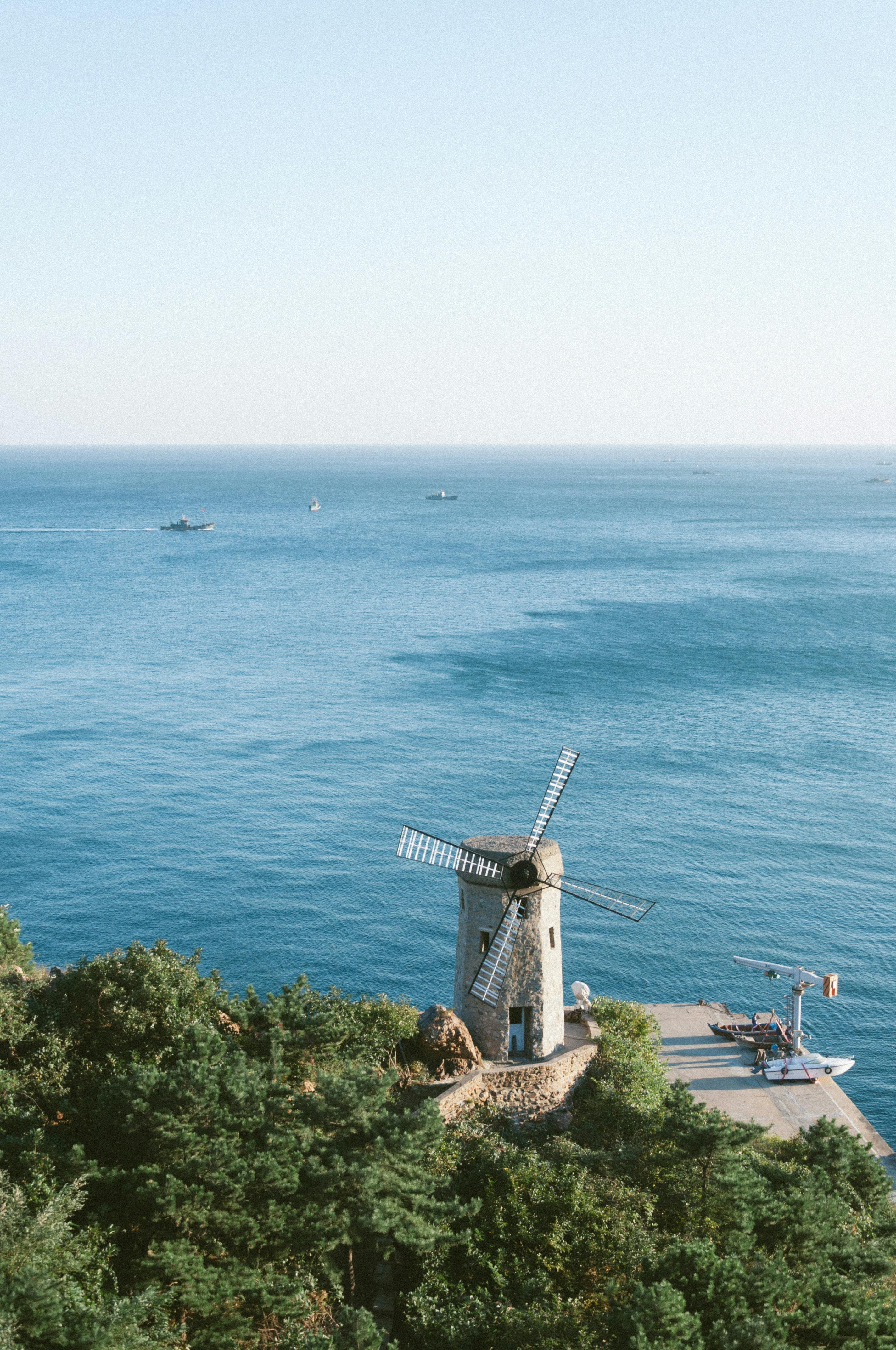 Picturesque view of a windmill by the sea in Dalian, China with greenery and clear skies.