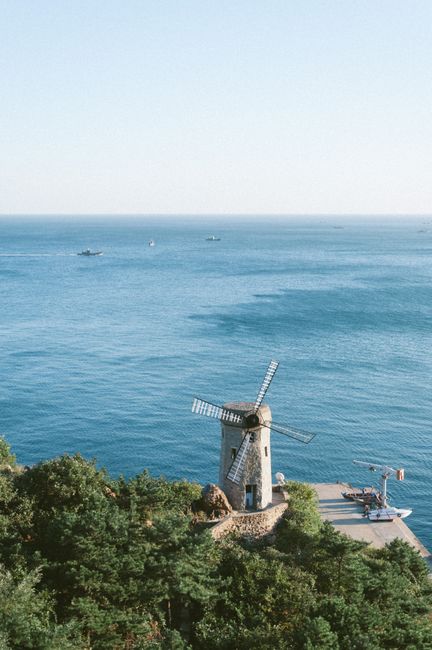 Picturesque view of a windmill by the sea in Dalian, China with greenery and clear skies.