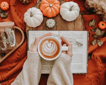 Top view of crop unrecognizable female with cup of latte on book sitting at wooden table with decorative pumpkins during autumn day
