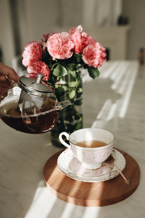 Free Crop unrecognizable person pouring hot tea from teapot into cup placed on table near bouquet of fresh roses in room Stock Photo