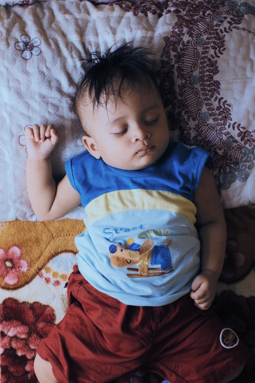 From above of calm ethnic little boy in casual clothes lying on comfortable bed and sleeping at home in daytime