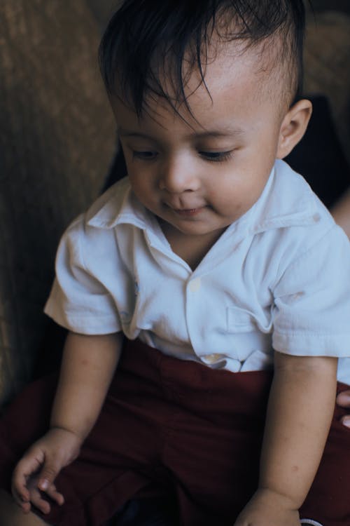 Crop calm little ethnic boy with dark hair in casual clothes sitting in room and looking down in daylight