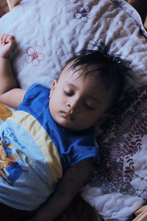 From above of tranquil little ethnic boy with dark hair in casual clothes lying on soft pillow on bed and sleeping in daytime