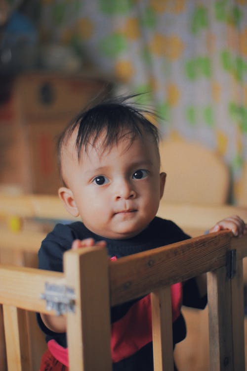 Ethnic boy standing on baby cot and looking at camera