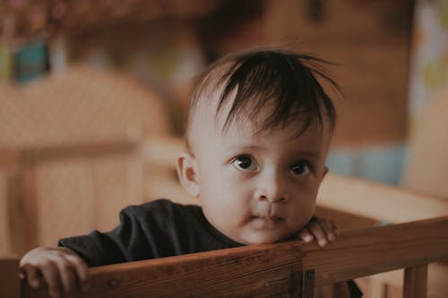 Adorable little ethnic baby with brown eyes and messy hair standing in crib and looking away with curiosity