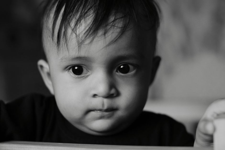 A close-up black-and-white portrait capturing the curious expression of a toddler indoors.