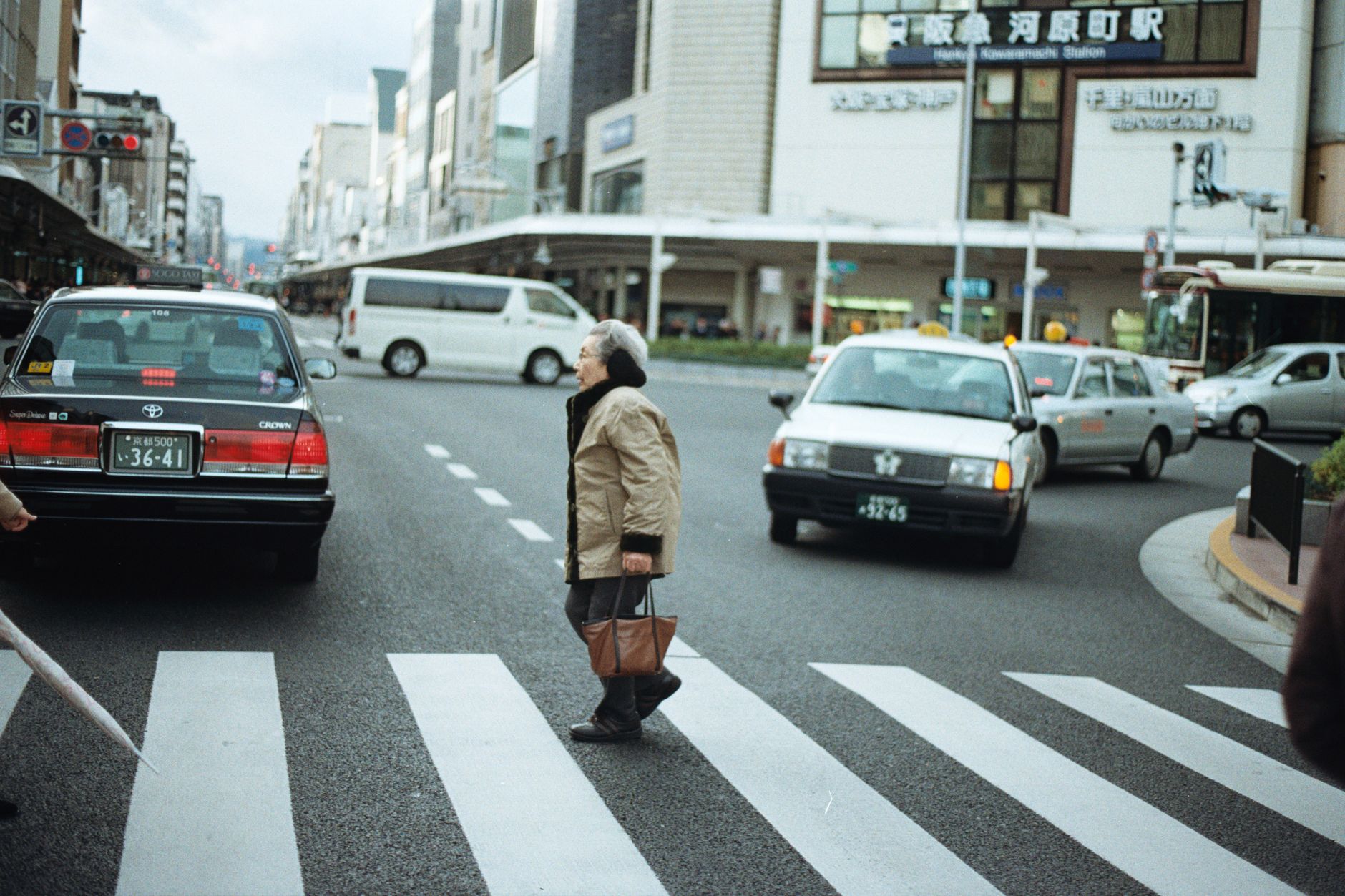 An elderly woman crosses a busy intersection in Osaka, Japan, surrounded by cars and city life.