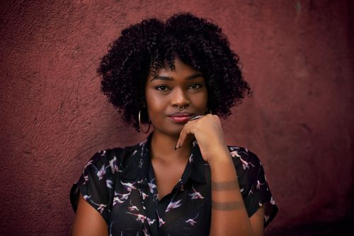 Free A woman with curly hair, wearing a floral top and hoop earrings, poses against a textured wall. Stock Photo