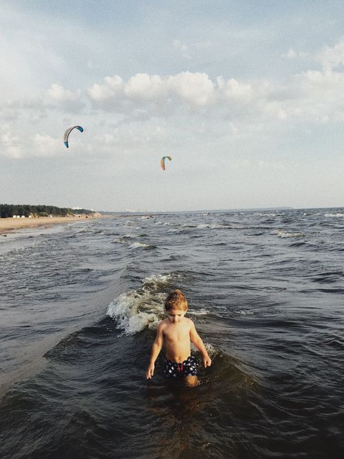 Free A young boy wades through the sea waves on a sunny beach, with colorful paragliders in the sky. Stock Photo