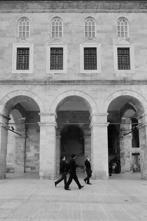Black and white side view of pedestrian people passing by stone historical building with arched columns and windows