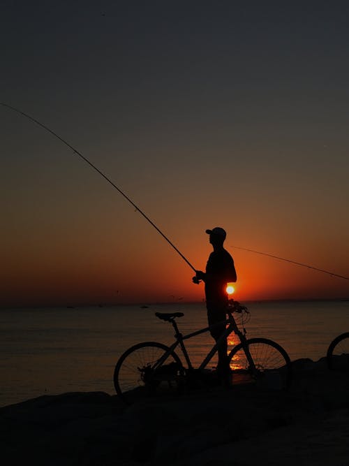 Free Silhouette of a Person Fishing on the Rocky Shore during Sunset  Stock Photo