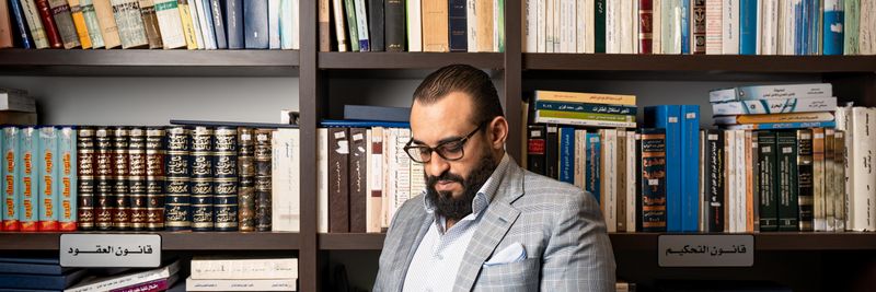 A bearded businessman in a suit reads a book in a library with shelves full of books.