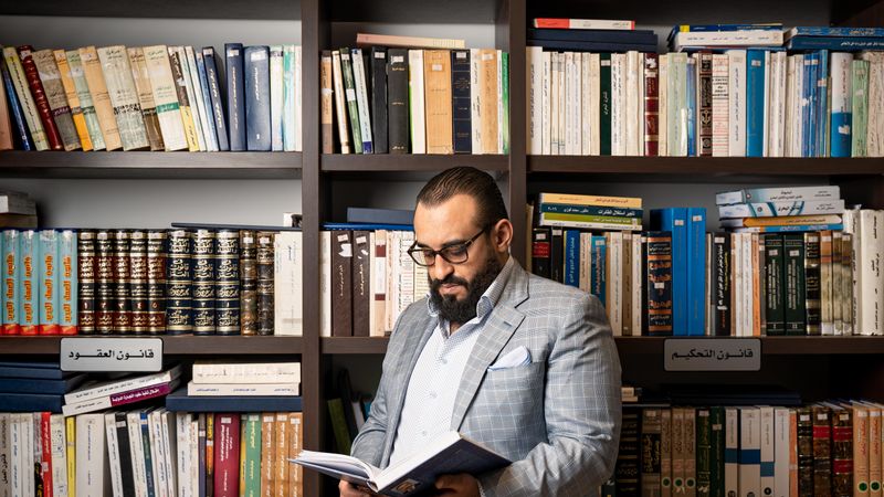 A bearded businessman in a suit reads a book in a library with shelves full of books.