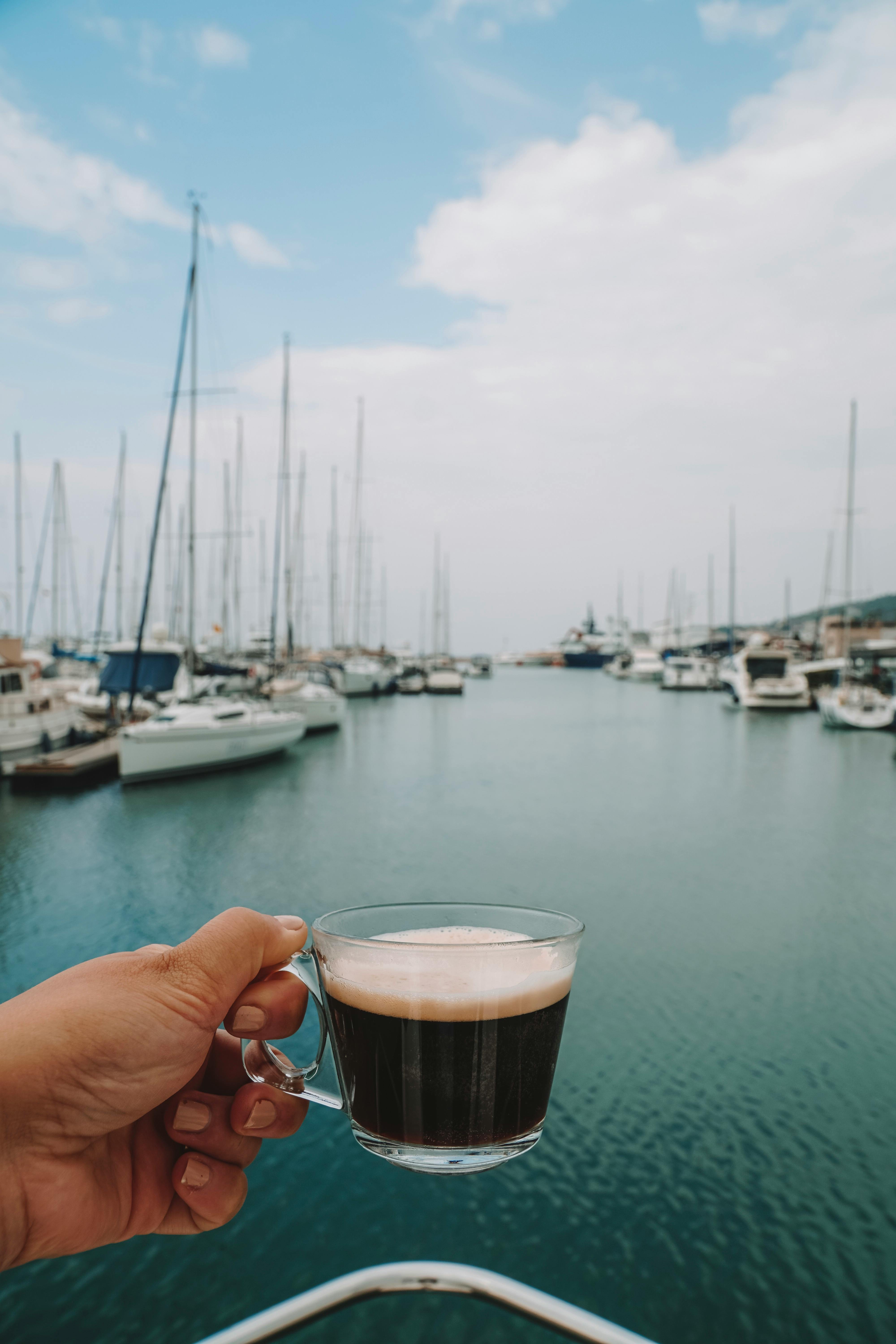 A hand holding coffee with Çeşme Marina's yachts in the background.