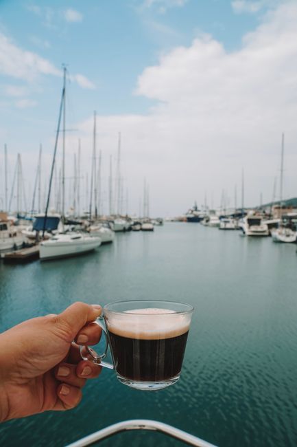 A hand holding coffee with Çeşme Marina's yachts in the background.
